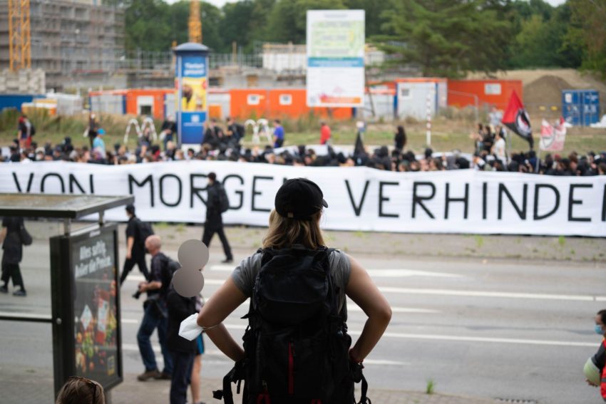 Im Vordergrund steht eine Frau mit Rucksack an einer Treppe, dahinter befindet sich die große Demonstration mit Bannern und Fahnen