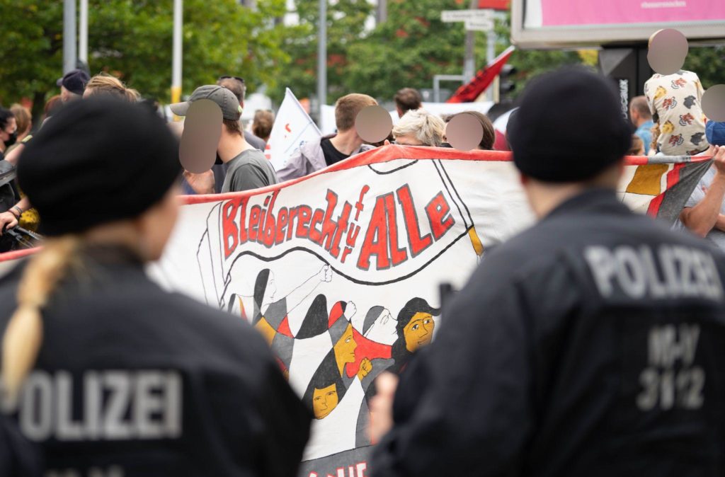 Ein Banner mit der Aufschrift "Bleiberecht für Alle", auf dem Personen auf einer Demonstration aufgemalt sind, davor stehen zwei Polizist*innen
