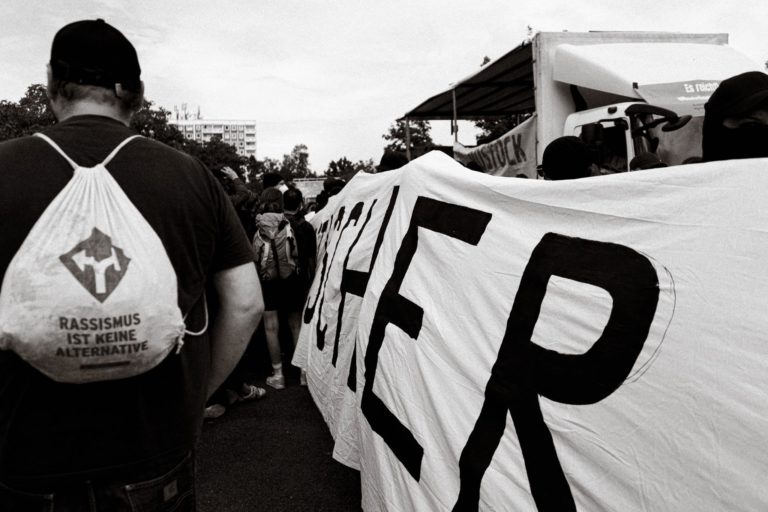 Ein Schwarzweißfoto von der Seitenansicht auf den schwarzen Block, links davon befindet sich eine Person mit einem Beutel, auf dem "Rassismus ist keine Alternative" abgedruckt ist